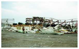 The aftermath of a Scud missile attack, showing a completely destroyed warehouse. A lone figure stands amidst the widespread debris and twisted metal structure.